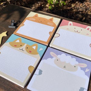 May include: Four square notepads featuring animal designs: a cat, a bunny, a sheep, and a corgi. Each notepad has a white grid pattern. The cat notepad is orange, the bunny is pink, the sheep is purple, and the corgi is blue.