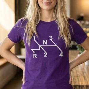 May include: A purple t-shirt with a white graphic of a car's gear shift pattern. The shirt displays the numbers 1, 2, 3, and 4, along with the letters "N" and "R" to indicate neutral and reverse. The shirt is short-sleeved.