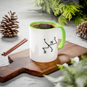 May include: White ceramic coffee mug with a green rim and handle. The mug has a black graphic of a car's gear shift pattern with the letters 'R', 'N', '1', '2', and '3'.