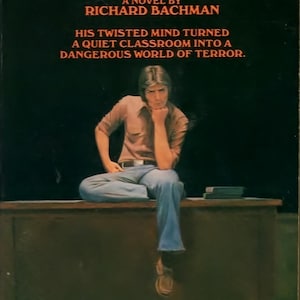 May include: Vintage paperback book cover for "Rage" by Richard Bachman. The cover displays the title in orange block letters, a seated figure, and text describing the novel's plot. The background is dark.