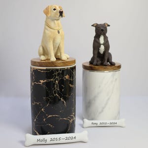 May include: Two ceramic pet urns with dog figurines on top. The urns are marble-patterned, one black with gold veins and the other white with gray veins.  A white bone-shaped plaque with the inscription "Molly 2015-2024" is in the foreground. Another white bone-shaped plaque with the inscription "Roxy 2012-2024" is in the background.