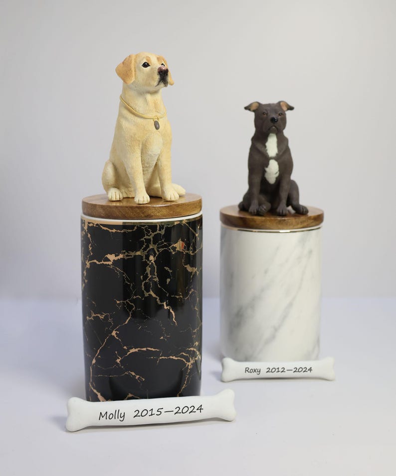 May include: Two ceramic pet urns with dog figurines on top. The urns are marble-patterned, one black with gold veins and the other white with gray veins.  A white bone-shaped plaque with the inscription "Molly 2015-2024" is in the foreground. Another white bone-shaped plaque with the inscription "Roxy 2012-2024" is in the background.