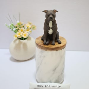 May include: A white marble urn with a wooden lid, featuring a ceramic figurine of a black and white pit bull dog sitting on top. A white bone with the inscription "Roxy 2012-2024" is in the foreground.