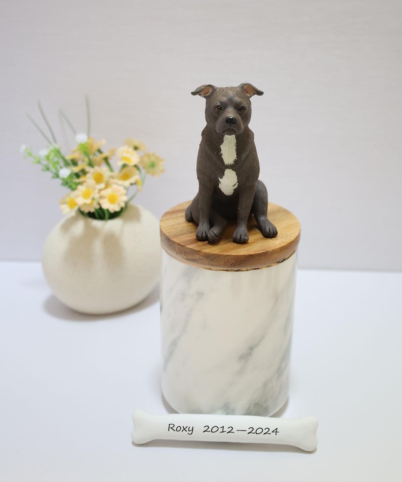 May include: A white marble urn with a wooden lid, featuring a ceramic figurine of a black and white pit bull dog sitting on top. A white bone with the inscription "Roxy 2012-2024" is in the foreground.
