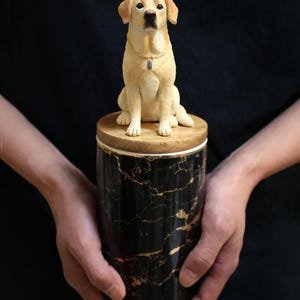 May include: A black and gold marble ceramic urn with a wooden lid. A small, realistic, cream-colored labrador retriever figurine sits on top of the lid.