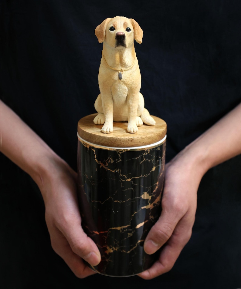 May include: A black and gold marble ceramic urn with a wooden lid. A small, realistic, cream-colored labrador retriever figurine sits on top of the lid.