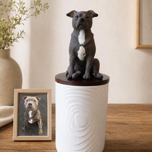 Puede incluir: Un recipiente cilíndrico de cerámica blanca con un patrón de ondas texturizado y una tapa de madera marrón oscuro. Una figura de perro gris y blanco se asienta sobre la tapa. Junto al recipiente hay un marco de madera con una foto de un perro.