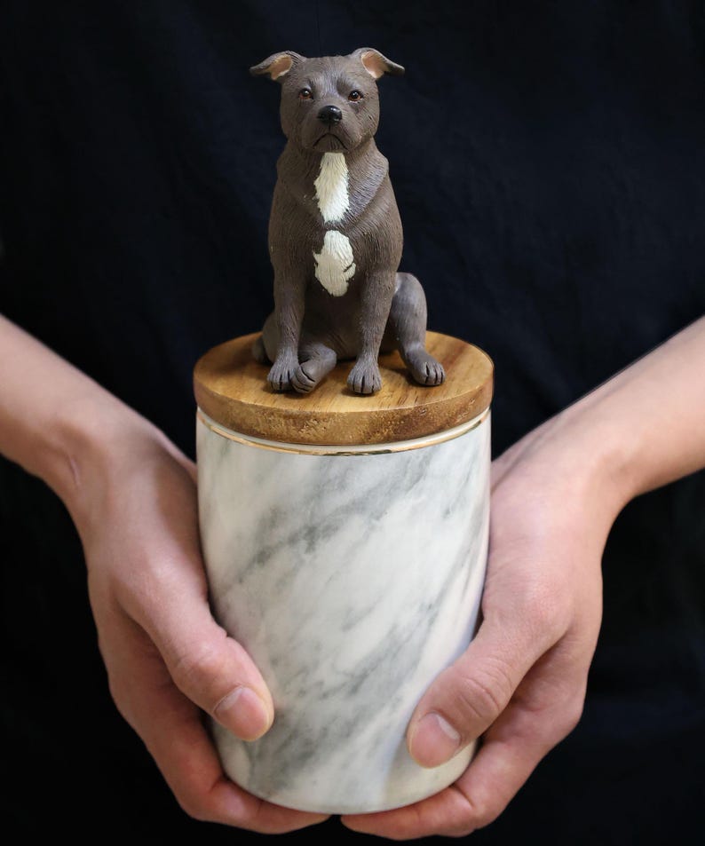 May include: A white marble urn with a wooden lid. A small, gray ceramic figurine of a pit bull terrier sits on top of the lid.