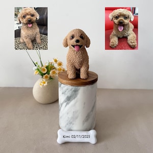 May include: A dog figurine with a smiling face is placed on a marble and wood container. The container has a white bone-shaped tag with the text "Kimi 02/11/2025". Two photos of a dog are in the background. A vase of flowers is to the left.
