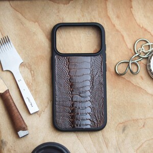 Può includere: Una custodia per telefono in pelle marrone con una cornice nera, con un motivo strutturato. Un cronometro argentato con una catena e strumenti per la lavorazione della pelle sono visibili su una superficie di legno.