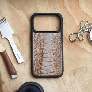 Può includere: Una custodia per telefono nera con un design marrone strutturato che ricorda la pelle di rettile. Un orologio da tasca argentato con catena e strumenti per la lavorazione della pelle sono su una superficie di legno.