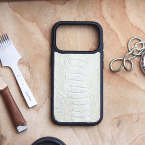 Può includere: Una custodia per telefono nera con un design strutturato color crema, forse in pelle di struzzo, è appoggiata su una superficie di legno. Un cronometro con catena e strumenti per la lavorazione della pelle sono anch'essi presenti. La custodia ha una cornice nera.