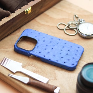 May include: A blue leather phone case with a textured surface and a pocket watch with a silver chain are displayed on a wooden surface. Leatherworking tools and a wooden watch box are also present.