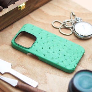 May include: A bright green phone case with a textured surface, a silver pocket watch, and a wooden box are arranged on a wooden surface. A leather crafting tool and a small bottle are also visible. The phone case has a black border.