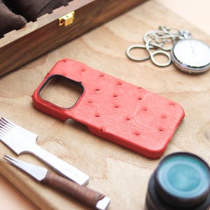 May include: A red leather phone case with a textured finish, placed with a silver pocket watch, leatherworking tools, and a wooden watch box. The items are on a wooden surface, indicating a focus on craftsmanship and accessories.