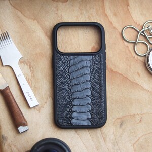 Può includere: Una custodia per telefono nera con un motivo in pelle di struzzo grigia è esposta su una superficie di legno. Sono presenti anche un cronometro argentato con catena e due strumenti per la lavorazione della pelle. Gli strumenti includono uno scalpello per cuciture e uno smussatore per bordi.