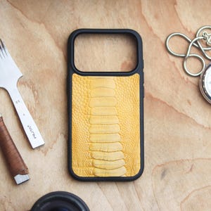 Può includere: Una custodia per telefono nera con retro in pelle di struzzo gialla. La custodia è circondata da strumenti per la lavorazione della pelle e un cronometro su una superficie di legno. La pelle ha un motivo strutturato a scaglie.