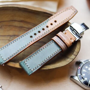 Può includere: Cinturino per orologio in due pezzi in pelle verde e beige. Il cinturino è cucito con un filo a contrasto e ha una fibbia in acciaio inossidabile. Il cinturino è esposto in una ciotola di legno, con un orologio e altri accessori sullo sfondo.