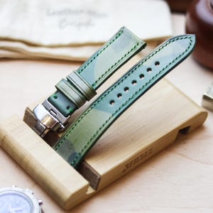 Könnte beinhalten: Grüne Camouflage-Uhrenarmbänder auf einem Holzständer. Die Armbänder haben eine Edelstahlschnalle. Eine Uhr mit schwarzem Armband ist zu sehen. Weitere Accessoires befinden sich im Hintergrund.