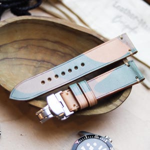 Könnte beinhalten: Armbänder für Uhren mit Tarnmuster und Edelstahlverschluss, präsentiert in einer Holzschale. Die Armbänder weisen eine Mischung aus Beige, Grün und Braun auf. Eine Uhr mit schwarzem Armband und eine weitere mit schwarzem Zifferblatt sind ebenfalls zu sehen.