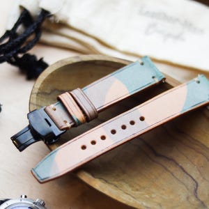 Peut inclure: Bracelet de montre à motif camouflage avec une base en cuir marron et des accents verts et beiges. Le bracelet est présenté avec un fermoir noir et un bol en bois. Une montre noire avec une lunette argentée est visible dans le coin.