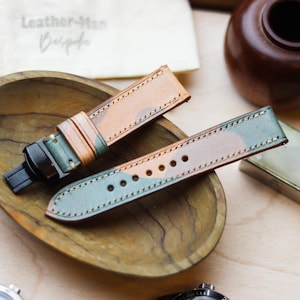 Könnte beinhalten: Zwei Leder-Armbänder mit einem Tarnmuster in Grün-, Beige- und Brauntönen werden in einer Holzschale präsentiert. Die Armbänder sind mit einem Kontrastfaden genäht. Der Text "Leather-Man Bespoke" ist im Hintergrund sichtbar.