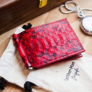 Op de afbeelding: Een rood en zwart geldclip-portemonnee met slangenleertextuur en een metalen clipmechanisme. De portemonnee rust op een wit stoffen trekkoordzakje met de tekst Leather-Man Bespoke in zwarte inkt op de voorzijde gedrukt.