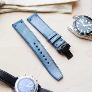 Può includere: Due cinturini per orologi in pelle blu chiaro con chiusure e cuciture nere. I cinturini sono accanto a un orologio argentato con quadrante nero e un orologio nero con quadrante blu.