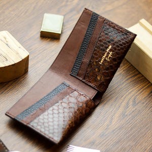 May include: Open brown leather wallet with a snakeskin pattern and black and blue accents. The wallet has the text "Leather-Man" in gold lettering. A gold lighter and wooden block are in the background.