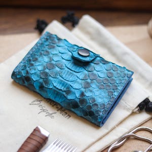 May include: A teal and black snakeskin-effect wallet with a press stud closure, displayed on a cream pouch. The wallet's distinctive scale pattern is prominent. A timer and tools are visible in the background, adding context to the scene.