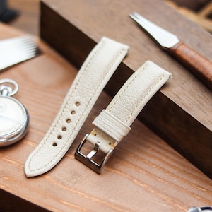 Pode incluir: Pulseiras de relógio de couro creme com fivela prateada, ao lado de um cronômetro, uma ferramenta envolta em couro e uma ferramenta de metal. As pulseiras de relógio são costuradas com uma linha contrastante. Os itens estão dispostos em uma superfície de madeira.