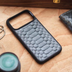 Può includere: Una custodia per telefono grigia con texture a pelle di serpente e bordo nero. Un portafoglio abbinato in pelle di serpente è visibile sullo sfondo, insieme a un cronometro e un piccolo oggetto rotondo verde. Gli oggetti sono disposti su una superficie di legno.