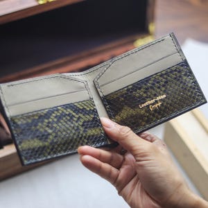 May include: A bi-fold wallet featuring a textured, olive green and black snakeskin pattern on the exterior card slots. The interior is lined with smooth, light beige leather. The text "Leather-Man" is embossed in gold foil on the right side.