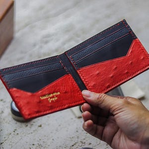 May include: A bi-fold wallet featuring a black interior with multiple card slots and a vibrant red exterior with a textured, dotted pattern. The wallet displays the gold-colored text Leather-Man Studio embossed on the lower left red panel.