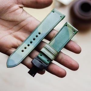 Può includere: Due cinturini per orologio con motivo mimetico verde e blu. I cinturini, con finitura strutturata, sono cuciti con filo verde. Uno ha fori di regolazione, l'altro una chiusura nera. Probabilmente in pelle.