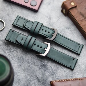 Può includere: Due cinturini per orologi in pelle verde scuro con fibbie argentate e nere. I cinturini sono cuciti con filo nero e hanno più fori per la regolazione. Lo sfondo include un taccuino in pelle e un telefono.