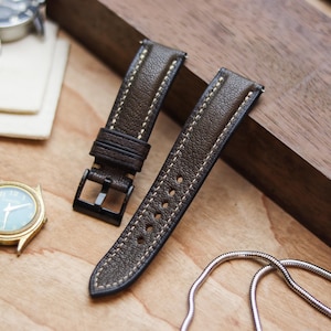 Pode incluir: Duas pulseiras de relógio de couro marrom com costura creme e fivelas pretas são exibidas em uma superfície de madeira. Um relógio dourado com mostrador azul claro e um relógio prateado também são visíveis, junto com uma corrente prateada.