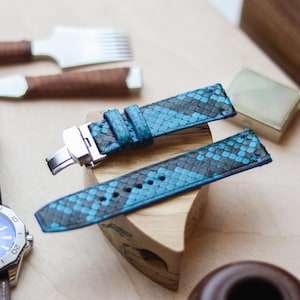 May include: Blue and black snakeskin watch band with a silver clasp, displayed on a wooden block. A watch with a blue face and brown leather band is visible in the background, along with leatherworking tools.