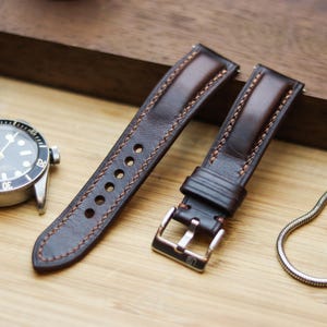 Könnte beinhalten: Nahaufnahme eines braunen Leder-Uhrenarmbands mit kupferfarbenen Nähten und einer silbernen Schnalle. Eine silberne Uhr mit schwarzem Zifferblatt und eine silberne Uhr mit weißem Zifferblatt sind ebenfalls im Bild. Die Artikel liegen auf einer Holzoberfläche.