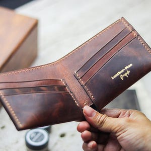May include: Open brown leather wallet with card slots and a bill compartment. The wallet has a distressed finish and visible stitching. The words "Leather-Man" and a signature are printed in gold on the interior.
