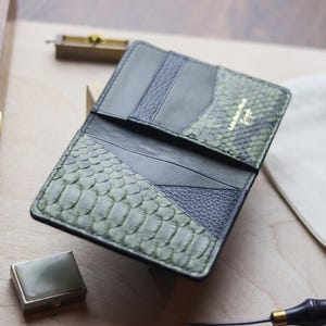 K&ouml;nnte beinhalten: Eine Geldb&ouml;rse aus einer Kombination von schwarzem Leder und texturierter olivgr&uuml;ner Schlangenhaut. Das Innere verf&uuml;gt &uuml;ber mehrere Kartenf&auml;cher und ein goldgepr&auml;gtes Logo mit der Aufschrift Leather-Hone Craft. Das Design umfasst glatte schwarze Lederpaneele neben Abschnitten aus genarbtem schwarzem Leder und gemusterten gr&uuml;nen Schuppen.