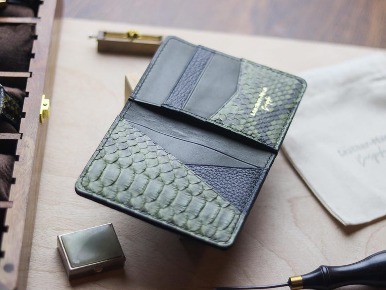K&ouml;nnte beinhalten: Eine Geldb&ouml;rse aus einer Kombination von schwarzem Leder und texturierter olivgr&uuml;ner Schlangenhaut. Das Innere verf&uuml;gt &uuml;ber mehrere Kartenf&auml;cher und ein goldgepr&auml;gtes Logo mit der Aufschrift Leather-Hone Craft. Das Design umfasst glatte schwarze Lederpaneele neben Abschnitten aus genarbtem schwarzem Leder und gemusterten gr&uuml;nen Schuppen.