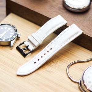 Può includere: Primo piano di un orologio e accessori. L'immagine presenta un orologio argentato con quadrante nero, un cinturino in pelle bianca con fibbia nera e un cronometro argentato. Il cinturino è in pelle bianca con cuciture bianche.