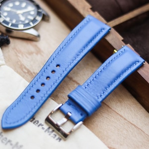 Pode incluir: Um par de pulseiras de relógio de couro azul brilhante com costuras brancas e uma fivela prateada. As pulseiras são exibidas em uma superfície de madeira, com um relógio ao fundo. As pulseiras são da Leather-Man.