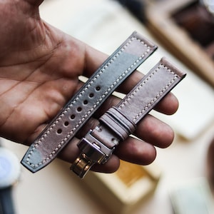 Può includere: Due cinturini per orologi in pelle con motivo mimetico nei toni del grigio, marrone e verde. I cinturini sono cuciti con filo bianco e tenuti in mano. Uno dei cinturini ha una fibbia argentata.