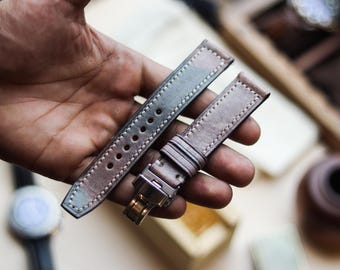 May include: A pair of leather watch straps in shades of gray, brown, and green, held in a hand. The straps have a camouflage pattern and are stitched with white thread. One strap has a silver buckle.