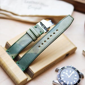 Könnte beinhalten: Ein grünes Camouflage-Leder-Uhrenarmband mit silberner Schnalle, ausgestellt auf einem Holzständer. Das Armband ist mit grünen Nähten versehen. Eine Uhr mit schwarzem Zifferblatt und silberner Lünette ist sichtbar.
