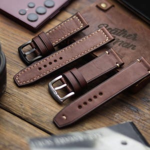 Könnte beinhalten: Nahaufnahme von mehreren braunen Lederarmbändern mit kontrastierenden weißen Nähten. Die Armbänder haben silberne und schwarze Schnallen. Im Hintergrund ein Lederetui mit dem Text "Leather Man" und ein Smartphone.