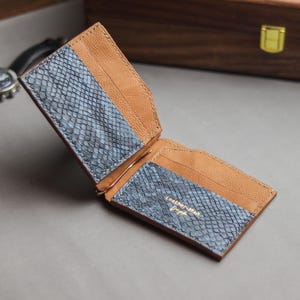 Op de afbeelding: Een bifold geldclip-portemonnee met bruin leer en blauwe panelen met slangenleertextuur. De binnenzijde bevat kaartsleuven en een centrale metalen geldclip. De tekst Leather-Man Craft is in goud gedrukt op het rechterblauwe paneel.