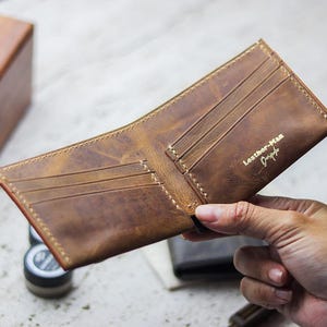 May include: Open brown leather wallet with card slots, held by a hand. The wallet has a distressed finish and the text "Leather-Man Design" in gold. The edges are stitched with a contrasting thread.